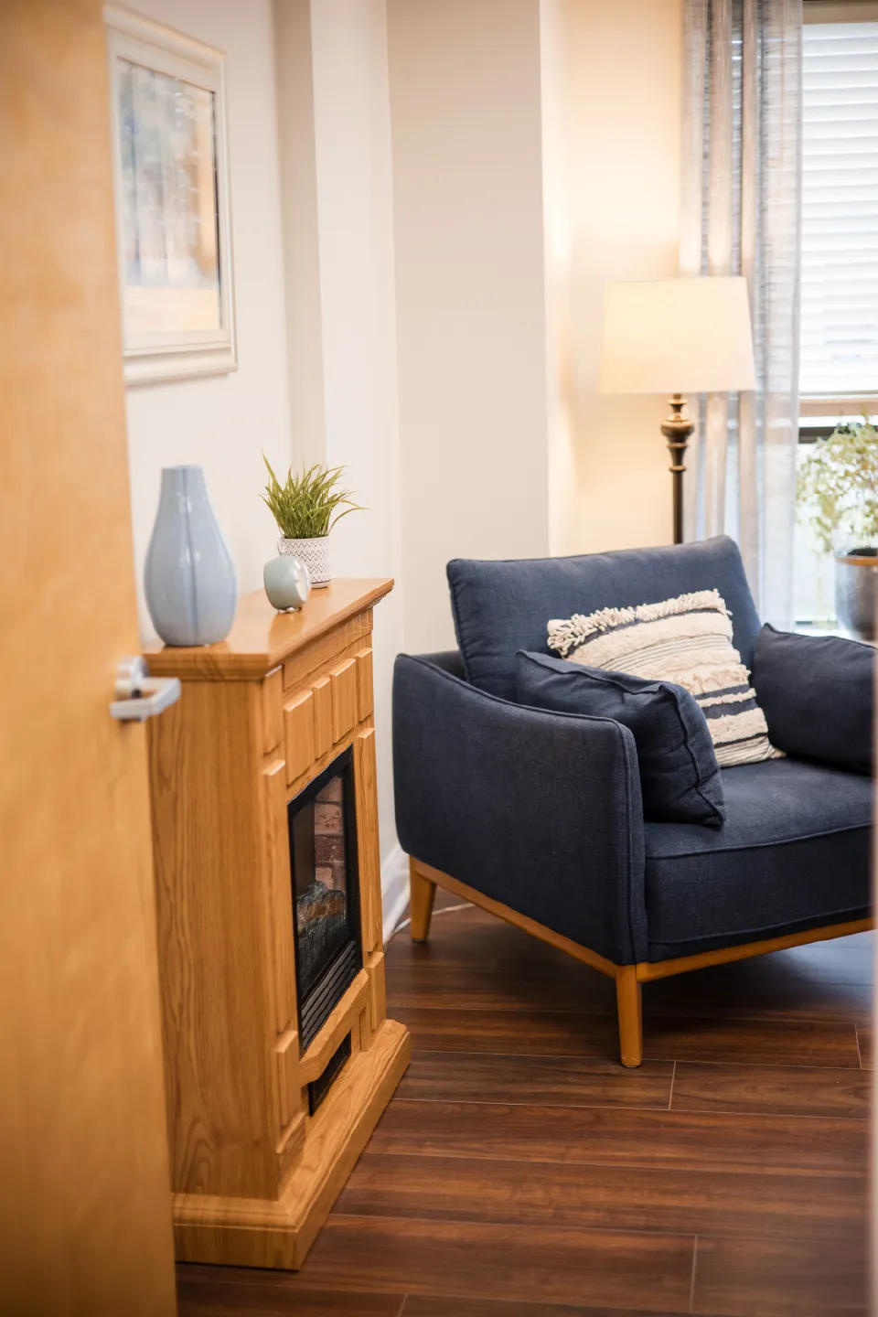 A cozy corner in the therapy practice with a blue armchair, textured pillow, wooden electric fireplace, and a floor lamp near window with sheer curtains.