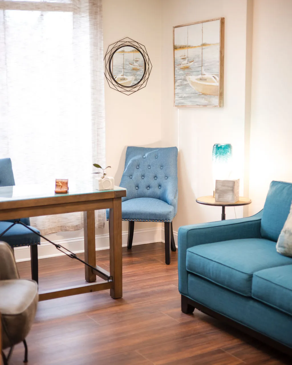 A cozy corner in the therapy practice with a blue armchair, textured pillow, wooden electric fireplace, and a floor lamp near window with sheer curtains.