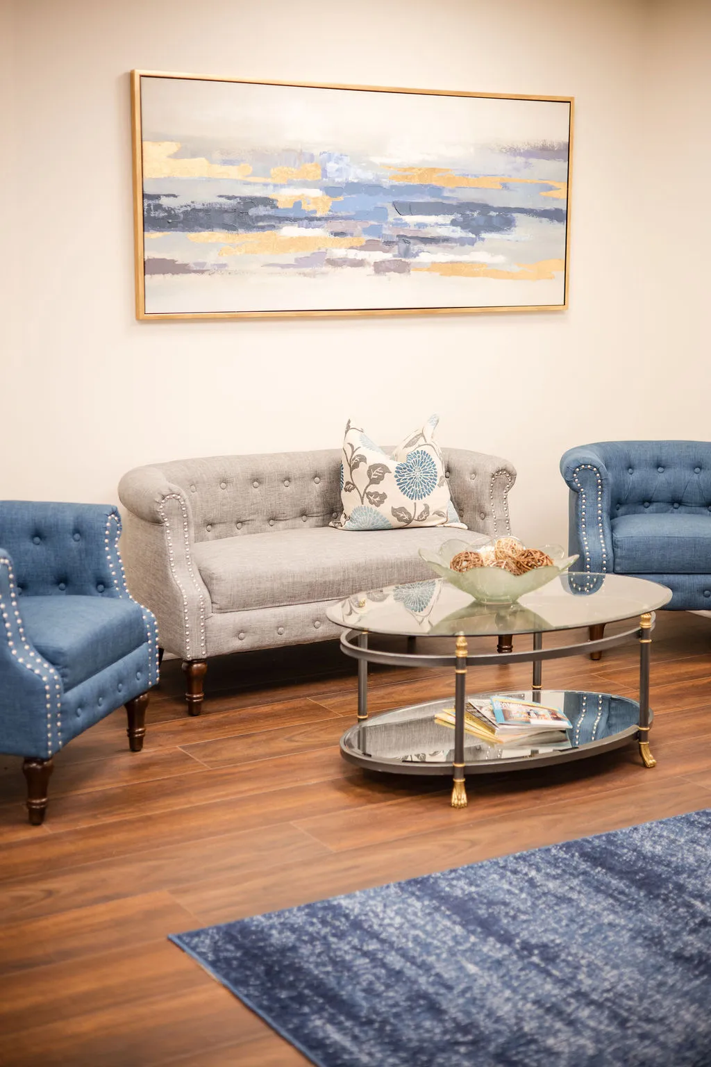 A seating area at the practice: a tufted gray sofa between two blue armchairs, a glass coffee table and an abstract painting above.