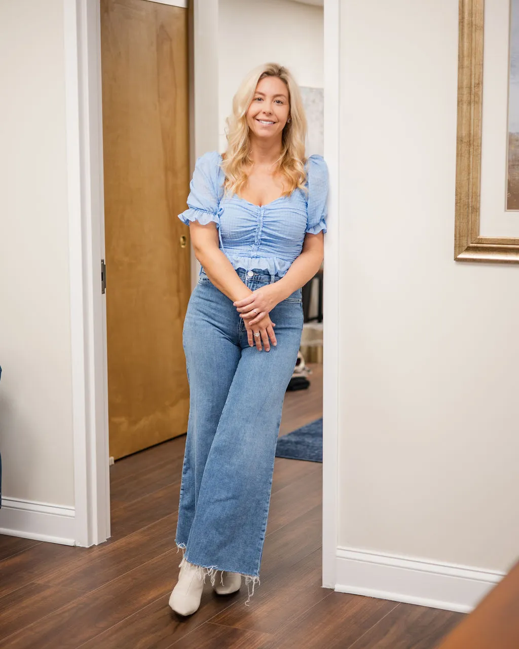 A smiling therapist in a light blue top and wide-leg jeans, leaning in a doorway at the practice.