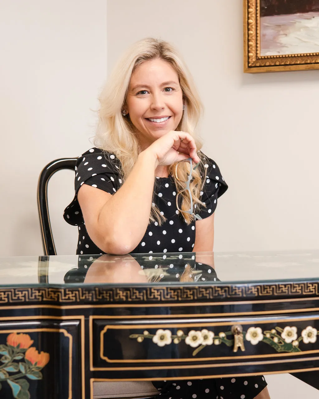 Lindsey Capelli, Living Well Counseling Center owner, in a black polka dot dress sitting at a decorative glass-top table holding eyeglasses.