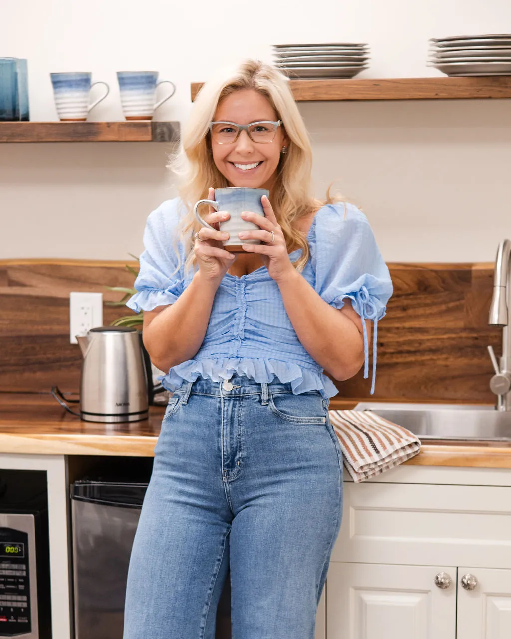 Smiling therapist with blonde hair and glasses holding a blue and white ceramic mug in the practice's kitchen.