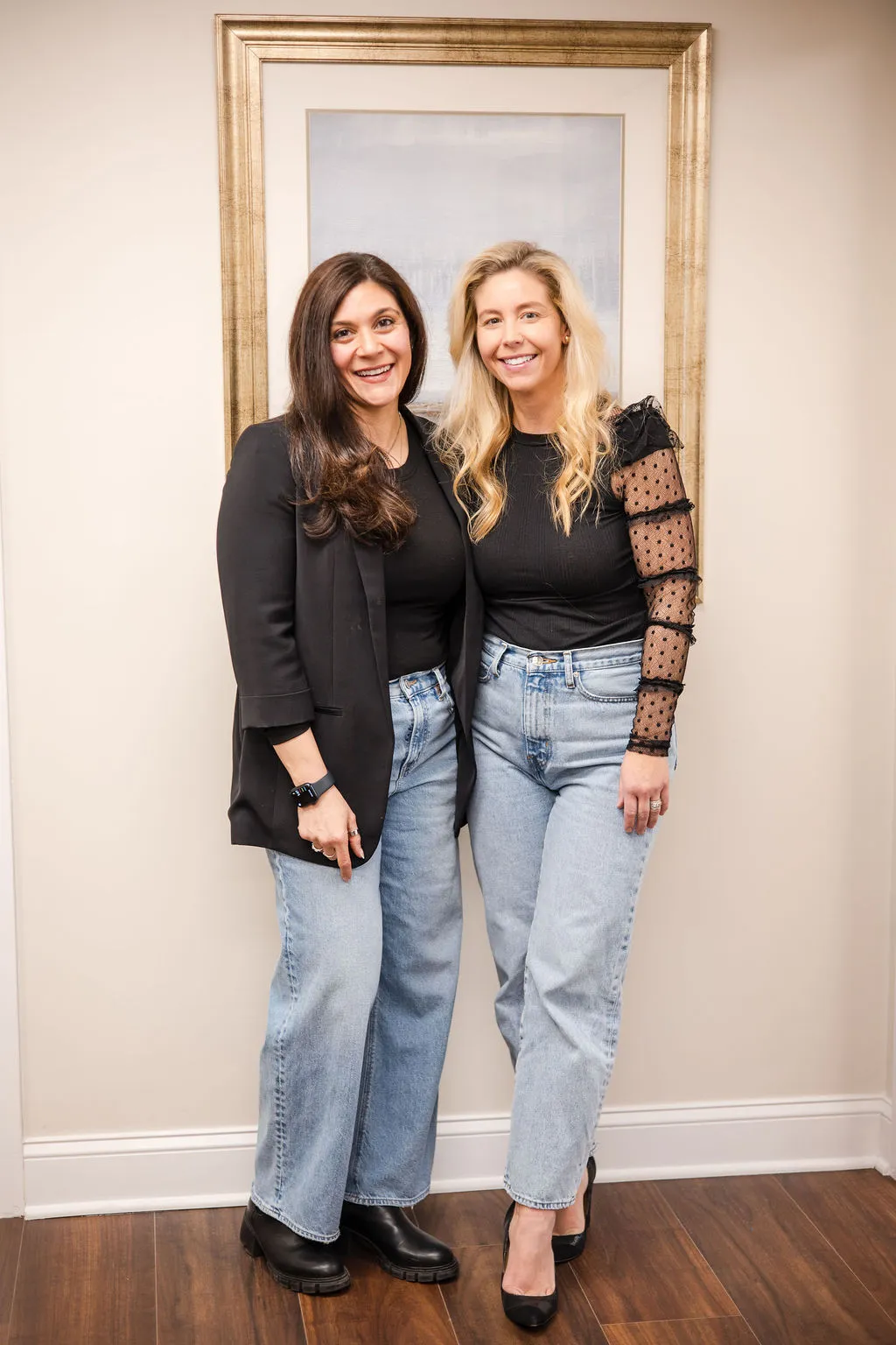 Two of the practice's therapists standing together, smiling, in black tops and jeans in front of a framed painting.