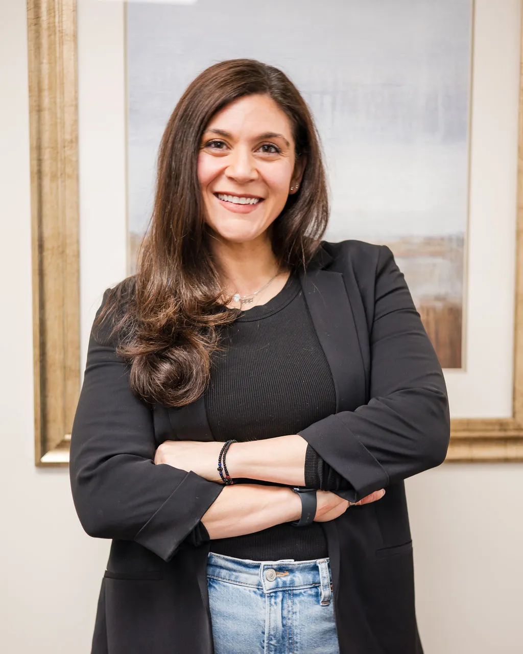 Marina Fried, therapist provider, with long brown hair wearing a black blazer, black top, and blue jeans standing with arms crossed in front of a framed abstract painting.